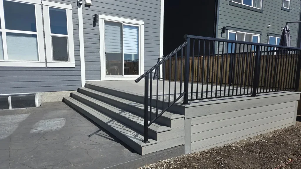 Gray wooden deck with stairs and black railing beside a house.
